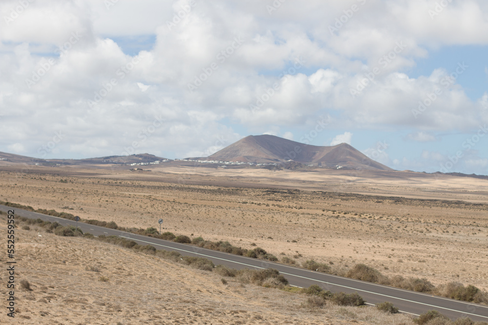 Desertic landscape. Lone black road runs through a desert plain. Mountains in the background. Blue sky with large white fluffy clouds. Yellow sand dunes with desert plants. Famara Beach, Lanzarote Desertic landscape. Lone black road runs through a desert plain. Mountains in the background. Blue sky with large white fluffy clouds. Yellow sand dunes with desert plants. Famara Beach, Lanzarote