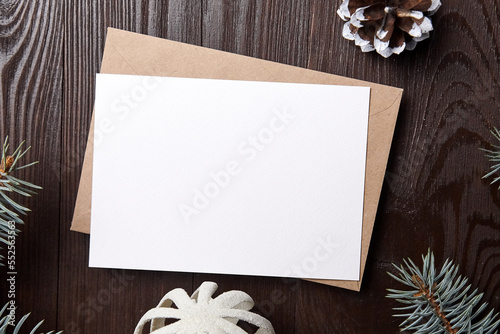 Christmas greeting card mockup with envelope, green fir tree branches and decorations on brown wooden background, top view, flat lay. White New Year holiday card with winter decor © mikeosphoto