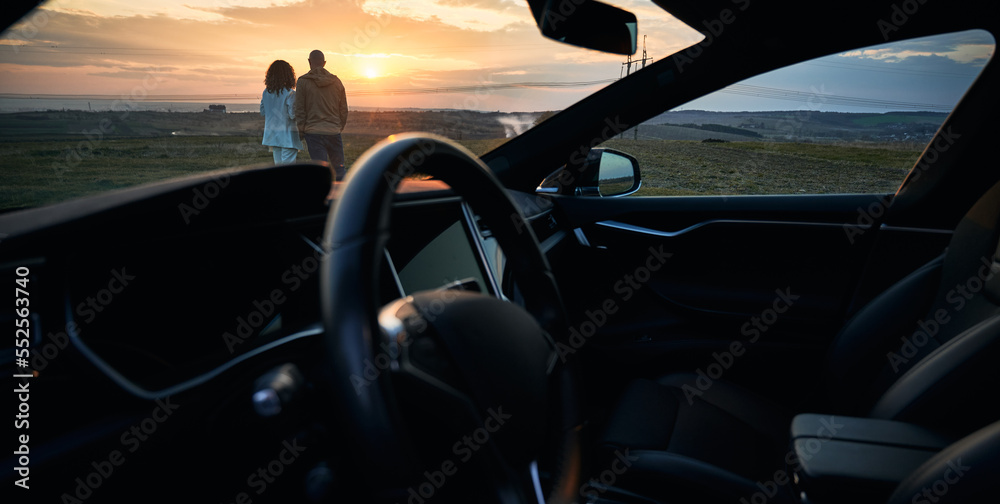 Beautiful view from automobile of young woman and man looking at orange evening sky during sunset. Snapshot of couple enjoying beauty of sundown with car steering wheel on foreground.