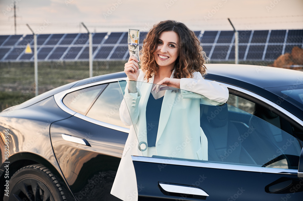 Portrait of business lady near open car, holding banknote against solar power plant. Female owner of ecological auto with electric motor demonstrating budget savings when using electric vehicles.