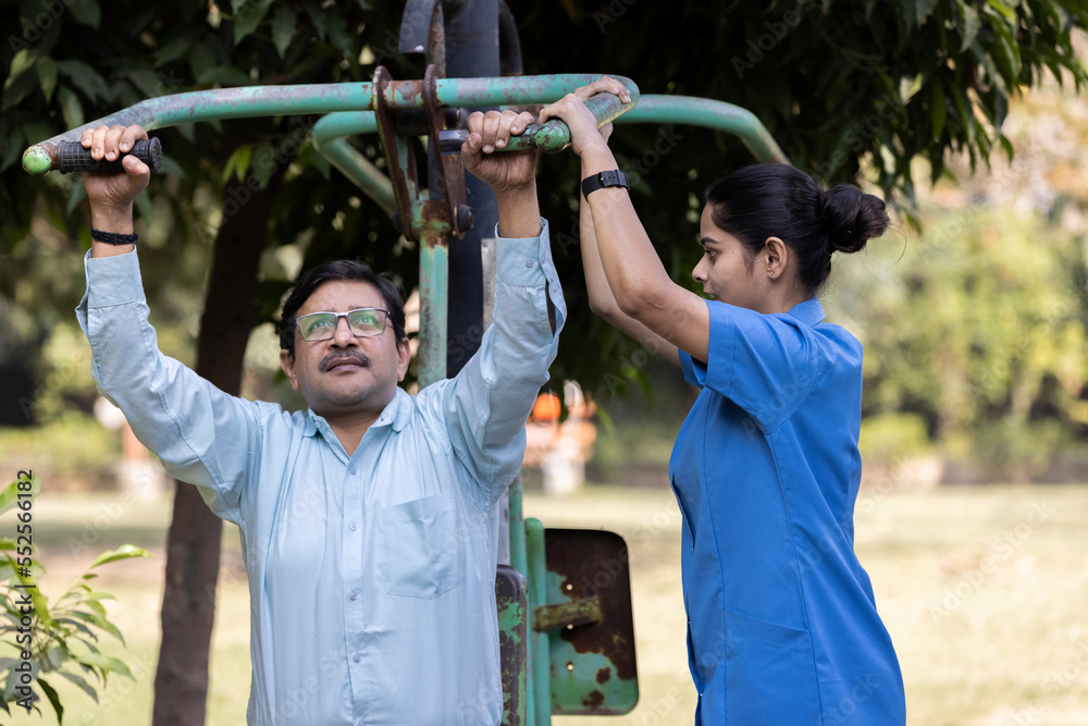 Obraz premium A physiotherapist or professional caregiver watches closely as a senior adult woman exercises on an outdoor gym.