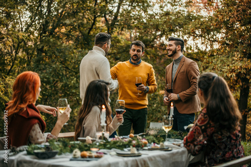 Group of elegantly dressed friends having a small boho dinner/wedding outdoors in a garden, drinking vine and talking to each other with a great mood and smiles.