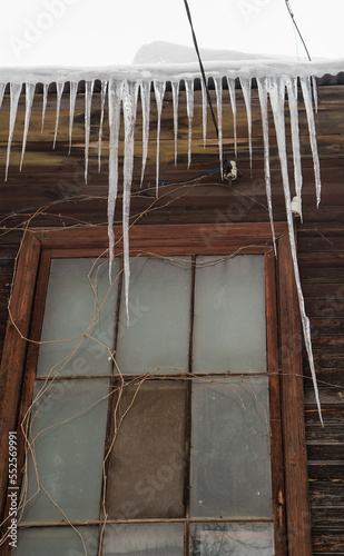Icy, dangerous icicles hang on the edge of the roof, winter or spring. Wall of an old wooden house with windows. Large cascades of icicles in smooth, beautiful rows. Cloudy winter day, soft light.