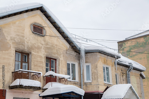 Snow-covered roof of an old house with windows, balconies and chimneys. There is snow and icicles on the roof, against the background of a gray sky. Cloudy, cold winter day, soft light.