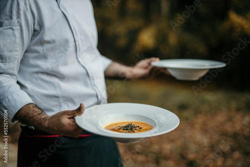 A male chef in a uniform brings a fresh prepared meal on plates to the table. A professional male chef is serving a wood on a boho dinner outdoors.