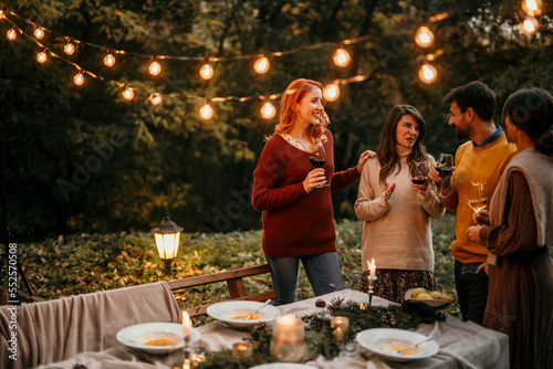 Group of elegantly dressed friends having a small boho dinner/wedding outdoors in a garden, drinking wine, and talking to each other with a great mood and smiles.
