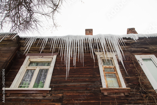Icy, large icicles hang on the edge of the roof, winter or spring. Plank wall of an old wooden house with windows. Large cascades of icicles in smooth, beautiful rows. Cloudy winter day, soft light.