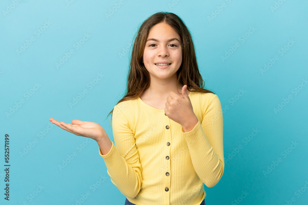 Little caucasian girl isolated on blue background holding copyspace imaginary on the palm to insert an ad and with thumbs up Little caucasian girl isolated on blue background holding copyspace imaginary on the palm to insert an ad and with thumbs up