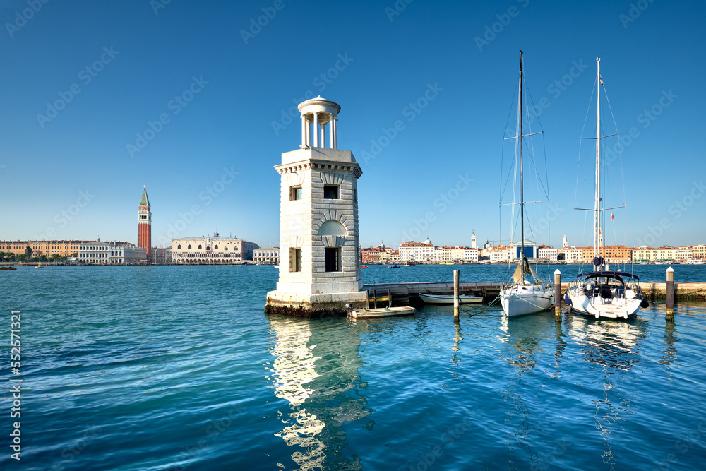 Marina and lighthouse tower on San Giorgio di Maggiore with city of Venice behind. Venezia, Italy, Europe. Sailing boats, yachts moored by pier. Sunshine, daylight, calm sea water with reflections.