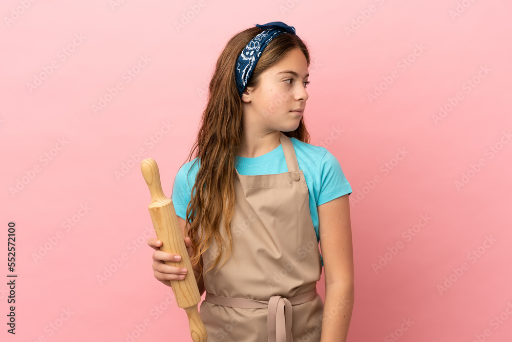 Little caucasian girl holding a rolling pin isolated on pink background looking to the side Little caucasian girl holding a rolling pin isolated on pink background looking to the side