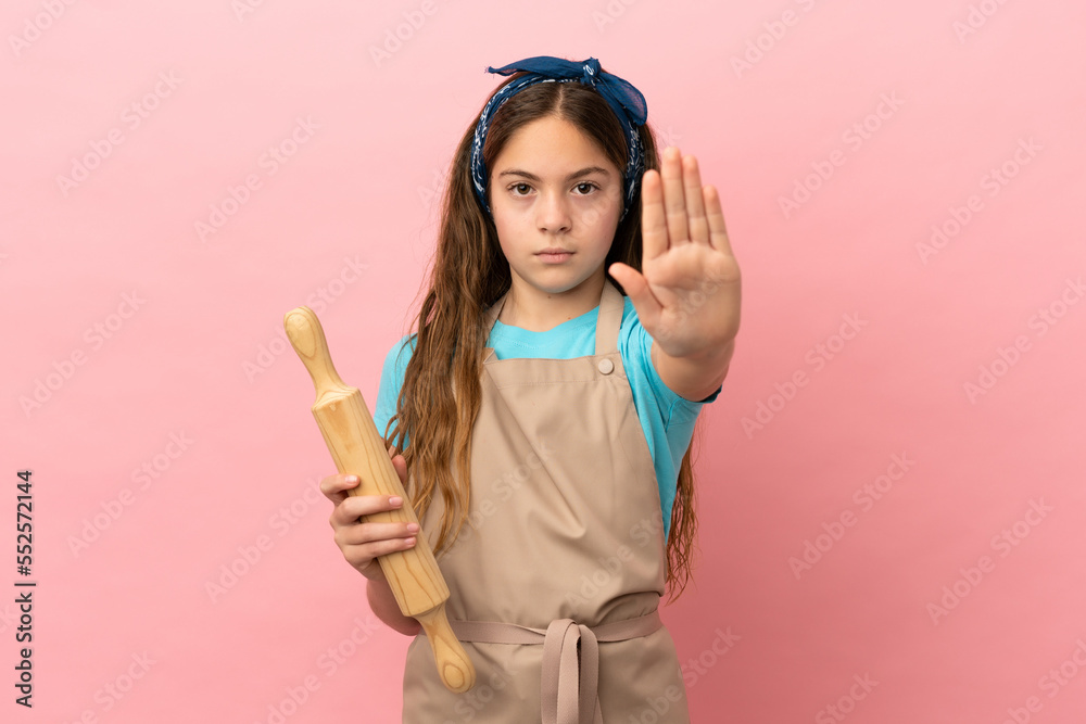 Little caucasian girl holding a rolling pin isolated on pink background making stop gesture Little caucasian girl holding a rolling pin isolated on pink background making stop gesture