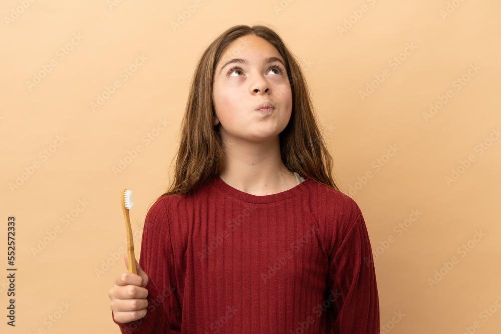 Little caucasian girl brushing her teeth isolated on beige background and looking up
