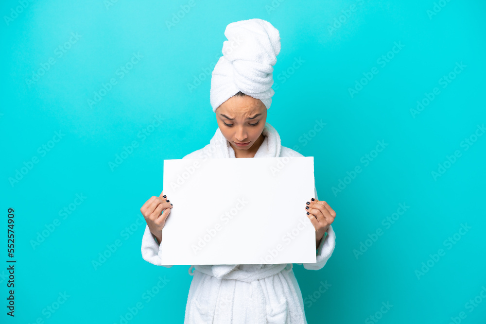 Young woman in a bathrobe with towel isolated on blue background holding an empty placard and looking it Young woman in a bathrobe with towel isolated on blue background holding an empty placard and looking it