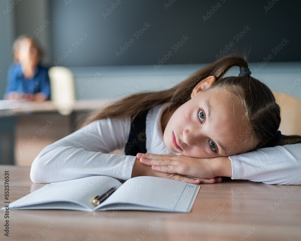Little caucasian girl is bored at the lesson at school. The schoolgirl folded her head on the desk and the teacher sits in the background. Little caucasian girl is bored at the lesson at school. The schoolgirl folded her head on the desk and the teacher sits in the background.