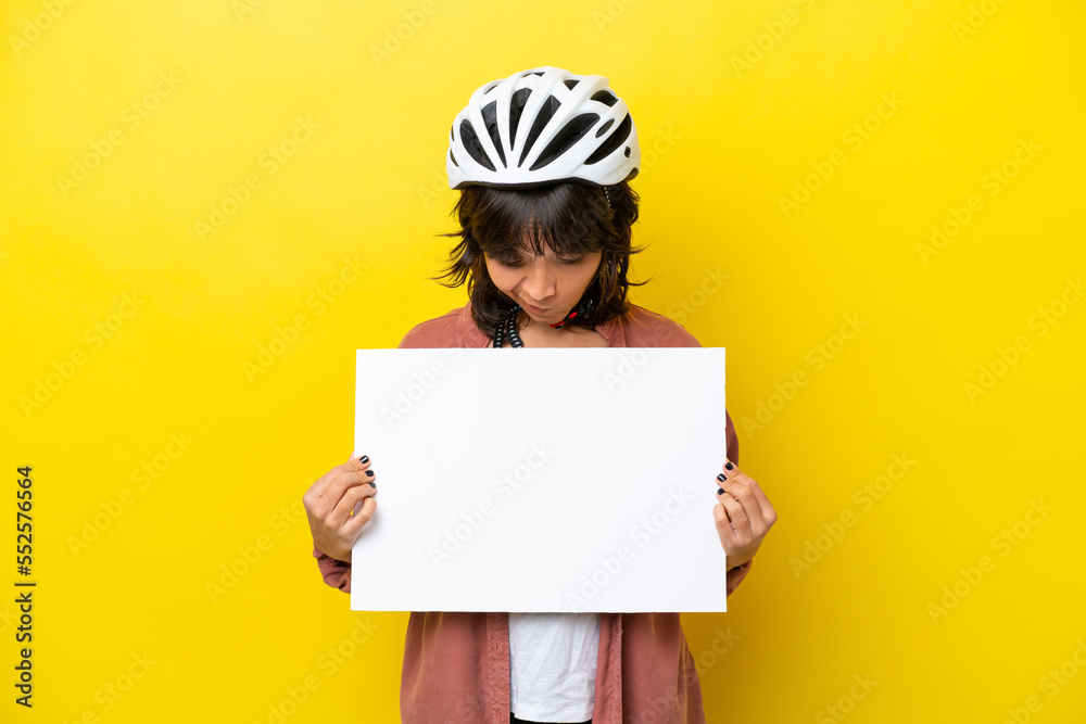 Young cyclist latin woman isolated on yellow background holding an empty placard and looking it Young cyclist latin woman isolated on yellow background holding an empty placard and looking it