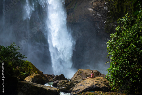 beautiful girl in pink bikini sits on rocks below eco chontales waterfall in Costa Rica; powerful waterfall in rainforest in Costa Rica