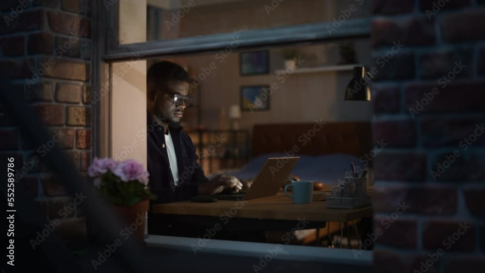 Portrait of Focused African American Man Working from Home on a Laptop, Being Productive at Night. Dedicated Black Male Web Developer Optimizing SAAS system, Thinking, Typing. View Through the Window