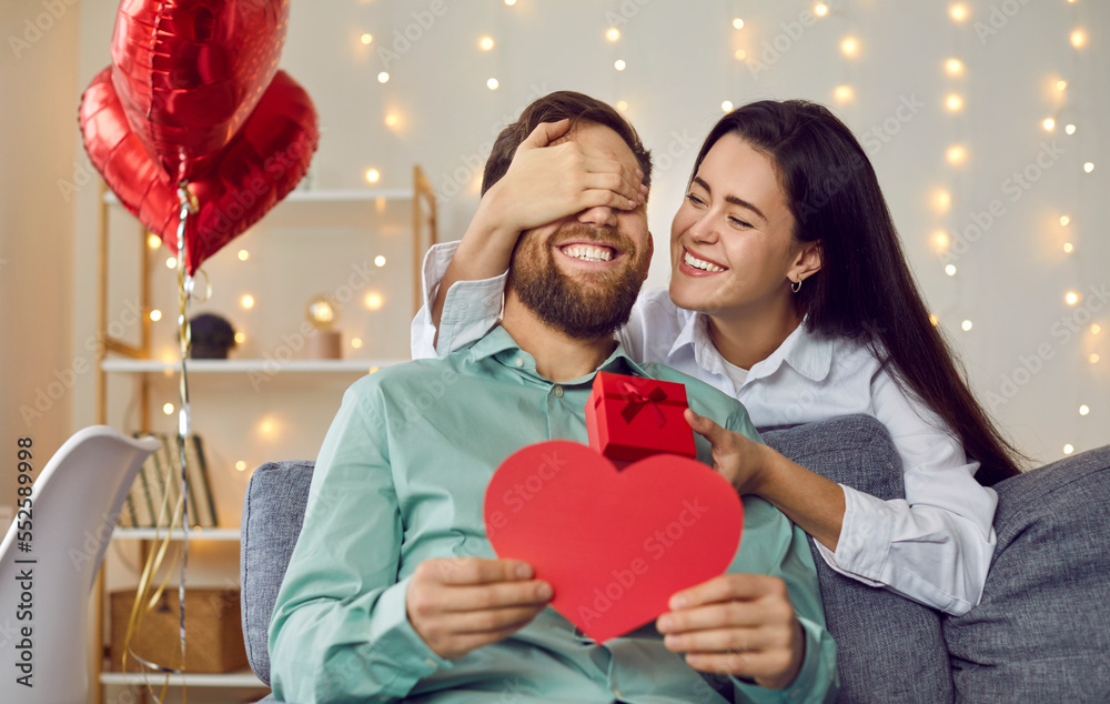 Joyful young couple celebrating St Valentine's Day. Loving woman makes  surprise for her boyfriend on Saint Valentine's Day. Happy girlfriend  covers man's eyes and gives him greeting card and gift box Photos