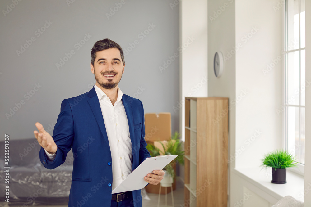 Portrait of professional realtor or real estate agent. Happy young man in suit standing in living room of new house for sale, holding clipboard, looking at camera and smiling Portrait of professional realtor or real estate agent. Happy young man in suit standing in living room of new house for sale, holding clipboard, looking at camera and smiling