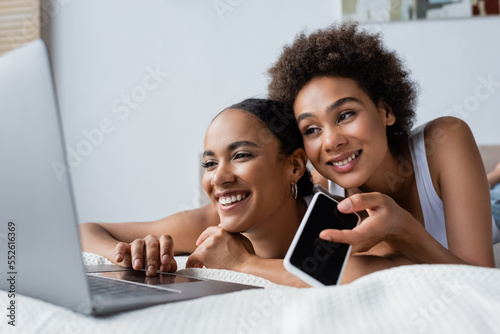 happy african american lesbian woman holding smartphone with blank screen while looking laptop near girlfriend.