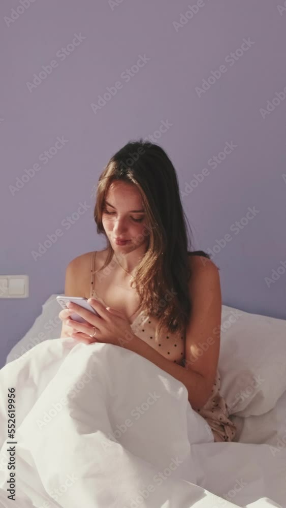 VERTICAL VIDEO, Happy woman texting on phone after waking up in the morning. Smiling girl having an online conversation, replying tap message while sitting in bed at home VERTICAL VIDEO, Happy woman texting on phone after waking up in the morning. Smiling girl having an online conversation, replying tap message while sitting in bed at home
