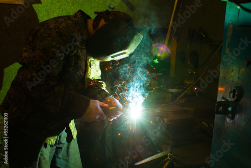 A masked worker performs spot welding work in his workshop, clamping a workpiece in a vise on a workbench