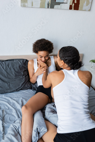 lesbian african american woman touching cheek of curly girlfriend in modern bedroom.