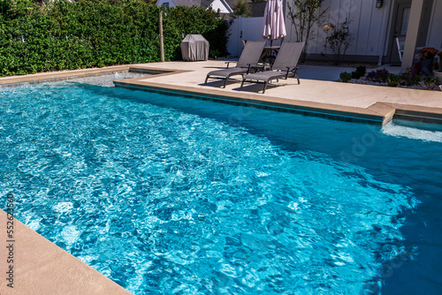 A rectangular new swimming pool with tan concrete edges in the fenced backyard of a new construction house