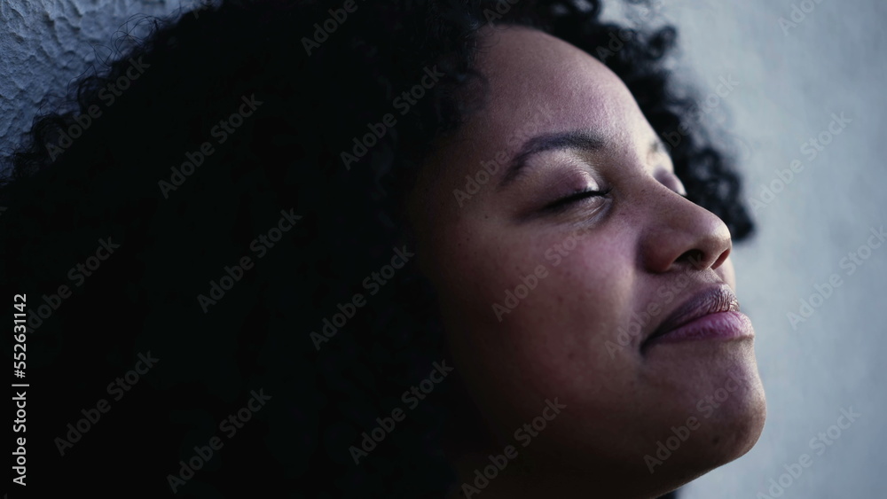 One hopeful meditative young black woman with eyes closed in contemplation. Faithful female person opening eye to sky smiling