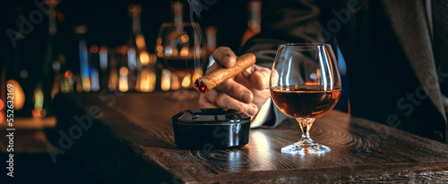 Man\'s hands with a cigar, elegant glass of brandy on the bar counter. Alcoholic drinks, cognac, whiskey, port, brandy, rum, scotch, bourbon. Vintage wooden table in a pub at night