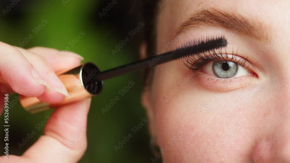Young woman painting eyelashes using black mascara half-face portrait. Close-up of painted female lashes of blue eye. Beauty, make-up and cosmetics concept.