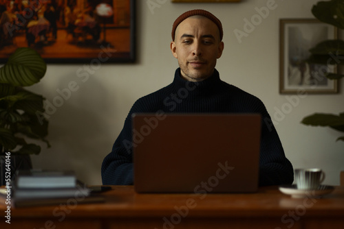 A caucasian man wearing a orange beanie and a blue knitted sweatshirt sitting at a wooden table working on a laptop.