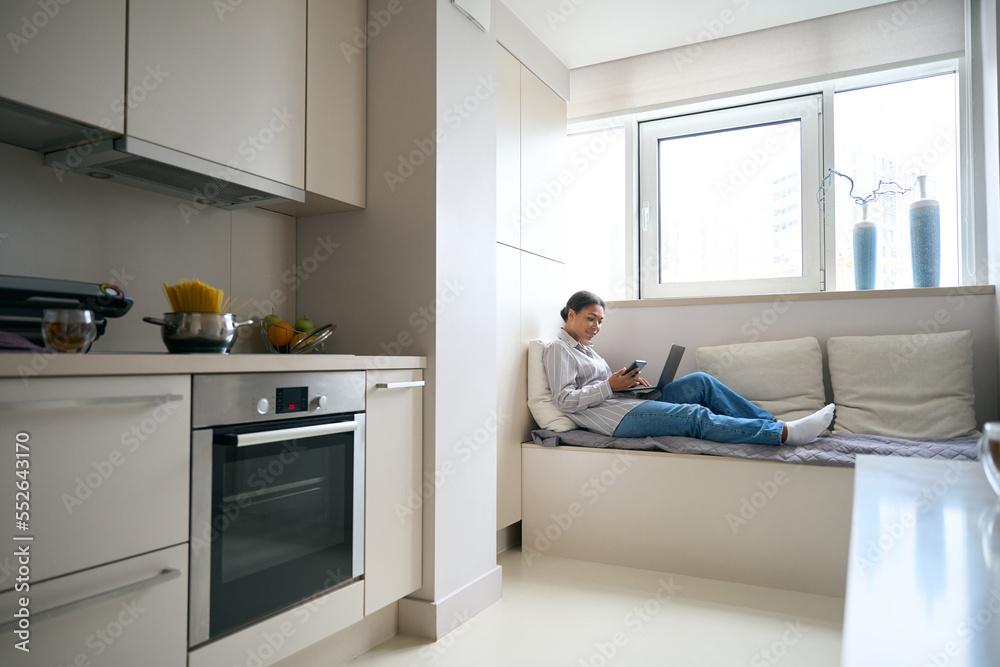 Pretty elegant female with smartphone and laptop is working online from home