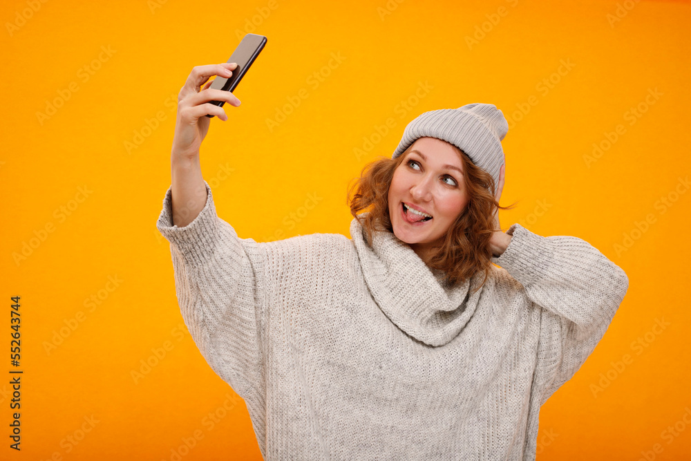 Young happy woman with curly hair in winter hat standing over yellow background making selfie. Happy New Year and Christmas concept Young happy woman with curly hair in winter hat standing over yellow background making selfie. Happy New Year and Christmas concept
