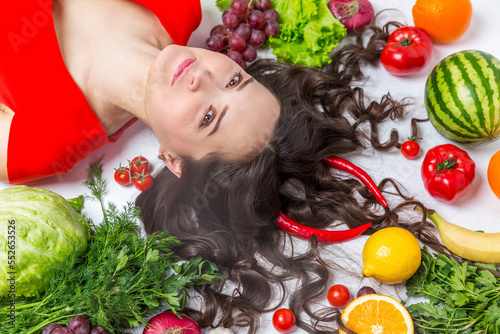 A young woman lies in fruits and vegetables on the floor on a white background. Pretty serious brunette in a red dress. Vegetarianism and healthy eating.