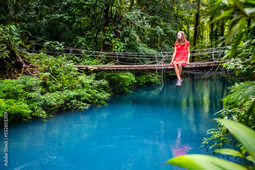 a girl in a red dress sits on a bridge over the blue rio celeste river in volcano tenorio national park; sky blue river surrounded by dense rainforest in costa rica