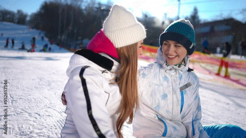 Rack focus from happy young woman hugging friend to smiling lady sitting on snow on winter resort. Satisfied Caucasian female friends enjoying leisure on vacations on sunny day outdoors Rack focus from happy young woman hugging friend to smiling lady sitting on snow on winter resort. Satisfied Caucasian female friends enjoying leisure on vacations on sunny day outdoors