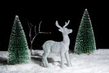 A toy shining silver deer standing in the snow in the forest among the Christmas trees at night on a black background