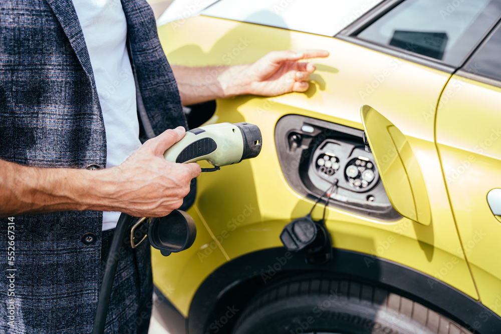 Fototapeta premium Close up of human hand is holding electric car charging before connect to yellow electric car outdoor.