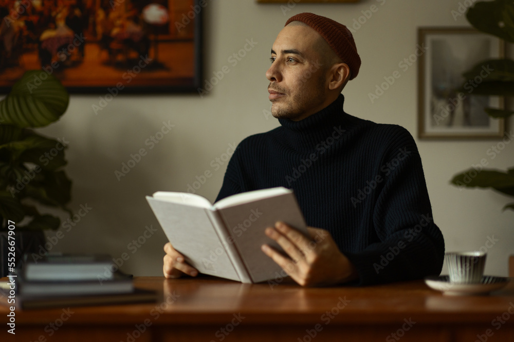 A caucasian man wearing a beanie and a knitted sweater sitting at a wooden table in a living room reading a book. A caucasian man wearing a beanie and a knitted sweater sitting at a wooden table in a living room reading a book.