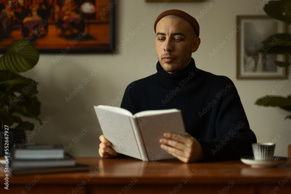 A caucasian man wearing a beanie and a knitted sweater sitting at a wooden table in a living room reading a book. A caucasian man wearing a beanie and a knitted sweater sitting at a wooden table in a living room reading a book.