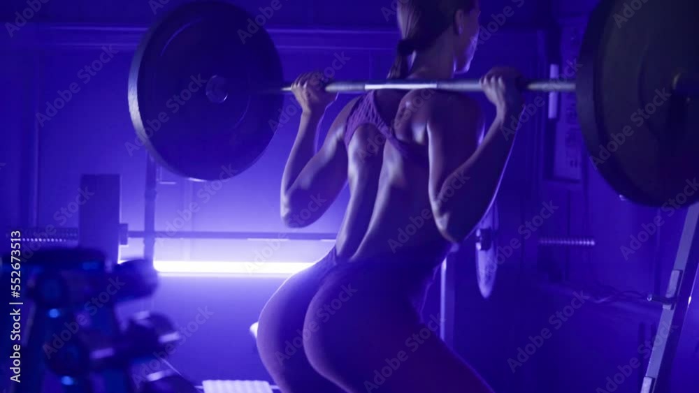 Back view of sporty fitness woman squatting with barbell in dark gym with blue light. Sportswoman bodybuilder in purple sportswear doing exercises for muscles of the buttocks and thighs. Close up.