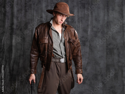 A young man in retro style, an adventure character. The hero of the adventure, a guy in a hat and a leather jacket, posing in the studio on a gray background