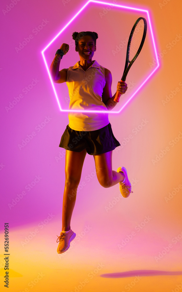 African american female player jumping and shaking fist by illuminated hexagon, copy space