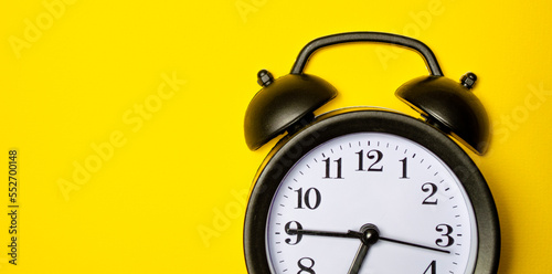 Small black alarm clock, black numbers, set the time placed on a table. Clock on isolated yellow background.