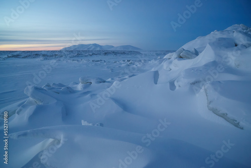 Winter Arctic landscape. Snow-covered ice hummocks near the sea coast. Tundra and mountains in the distance. Twilight in the afternoon in December in the Arctic. The cold climate of the polar region.