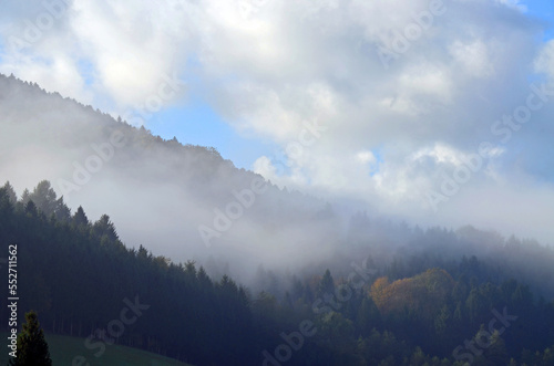 Niedrige Wolke schmiegt sich an einen bewaldeten Berg. Der Hintergrund ist neblig, der Himmel blau. 