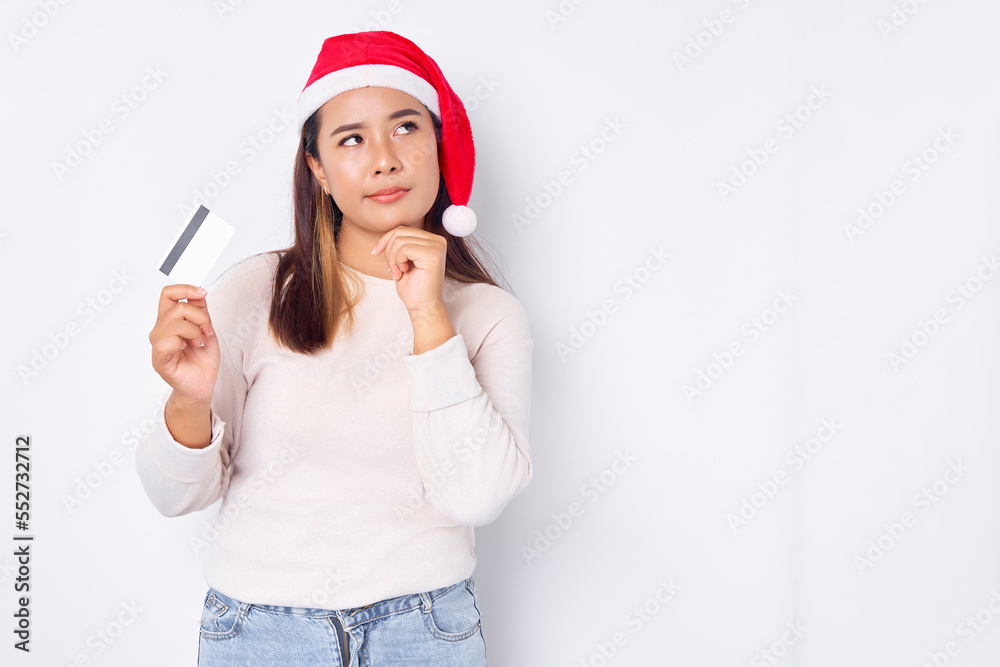 Pensive young Asian woman in a Christmas hat showing blank white credit card and touching chin on hand isolated on white background Pensive young Asian woman in a Christmas hat showing blank white credit card and touching chin on hand isolated on white background