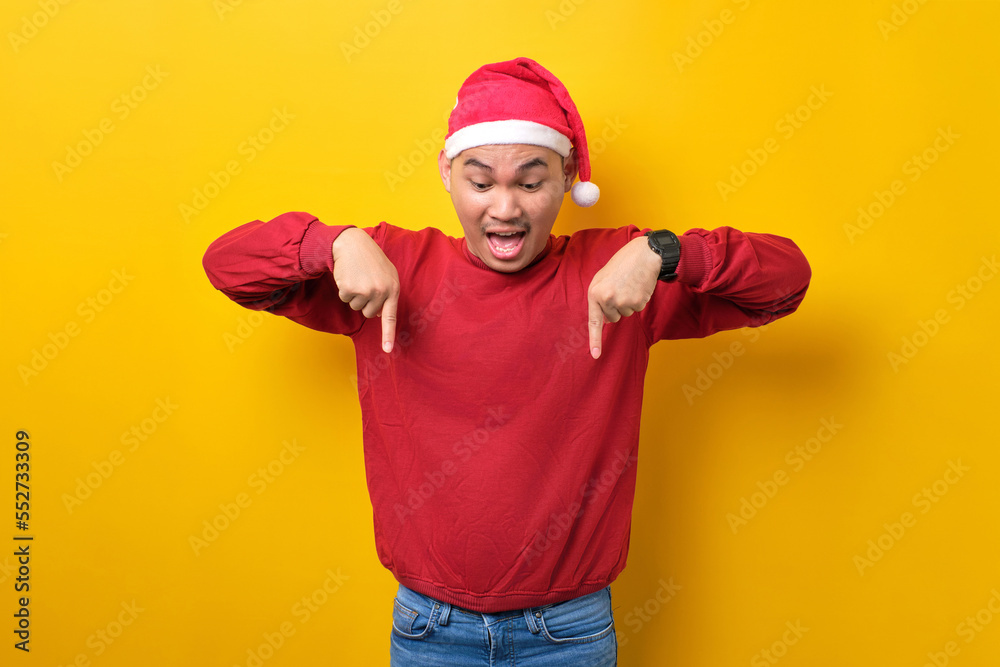 Naklejka premium Surprised young Asian man in Santa hat pointing fingers down, demonstrating free copy space for advertisement on yellow studio background. celebration Christmas holiday and New Year concept