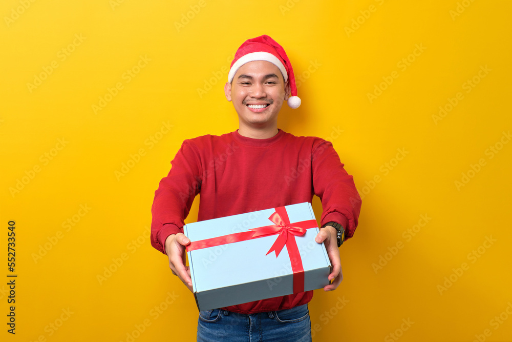 Smiling young Asian man in Santa hat holding gift box with ribbon on yellow studio background. celebration Christmas holiday and New Year concept Smiling young Asian man in Santa hat holding gift box with ribbon on yellow studio background. celebration Christmas holiday and New Year concept